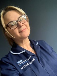 Jody, lady with blonde hair tied back in a ponytail. Wearing glasses and smiling. Wearing a dark blue nurses uniform