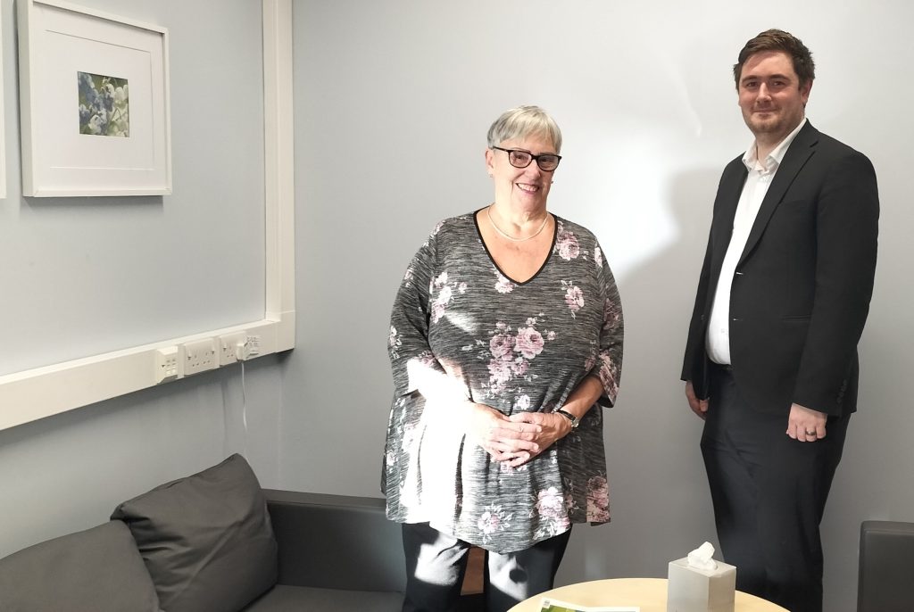 Margaret, short grey hair with glasses, stood with the Middlesbrough Mayor, Chris Cook, in a black suit in the quiet room, which has grey sofas and a table with pictures on the wall.